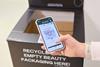 Marks & Spencer in-store beauty recycling bin behind customer holding a phone showing offers