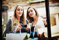 Two women looking at products in a shop window and taking a photo with their phone