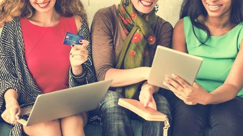 Three women sat next to each other looking at laptops and tablets and one holding a credit card