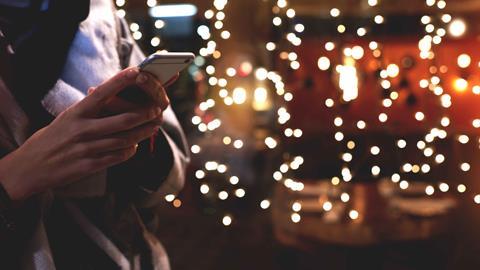 Woman-holding-mobile-phone-against-background-of-Christmas-lights