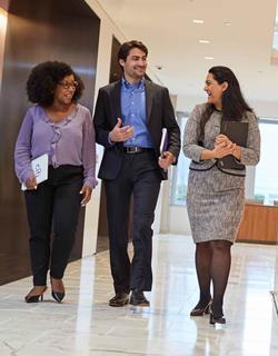 Three people talking and walking in an office environment, two women holding clipboards (one black, one Asian) and one white man in the middle
