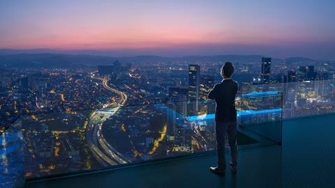 A man looks out from a balcony at a city by night, with the sun setting
