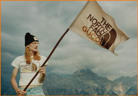 Man outdoors against mountainous background in a T-shirt that reads 'The North Face Gucci' and waving a flag with the same caption