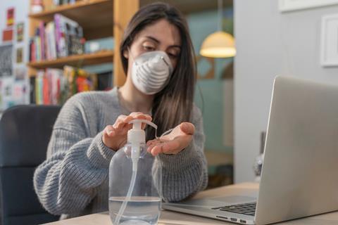Woman working on laptop at home wearing mask and sanitising her hands