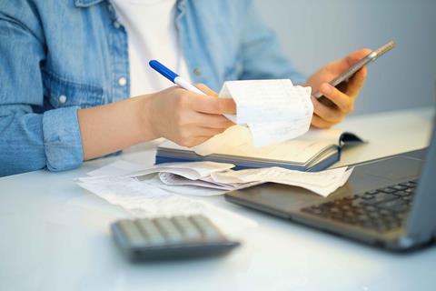 Woman balancing finances with phone, calculator, receipts and laptop, shown from shoulders down